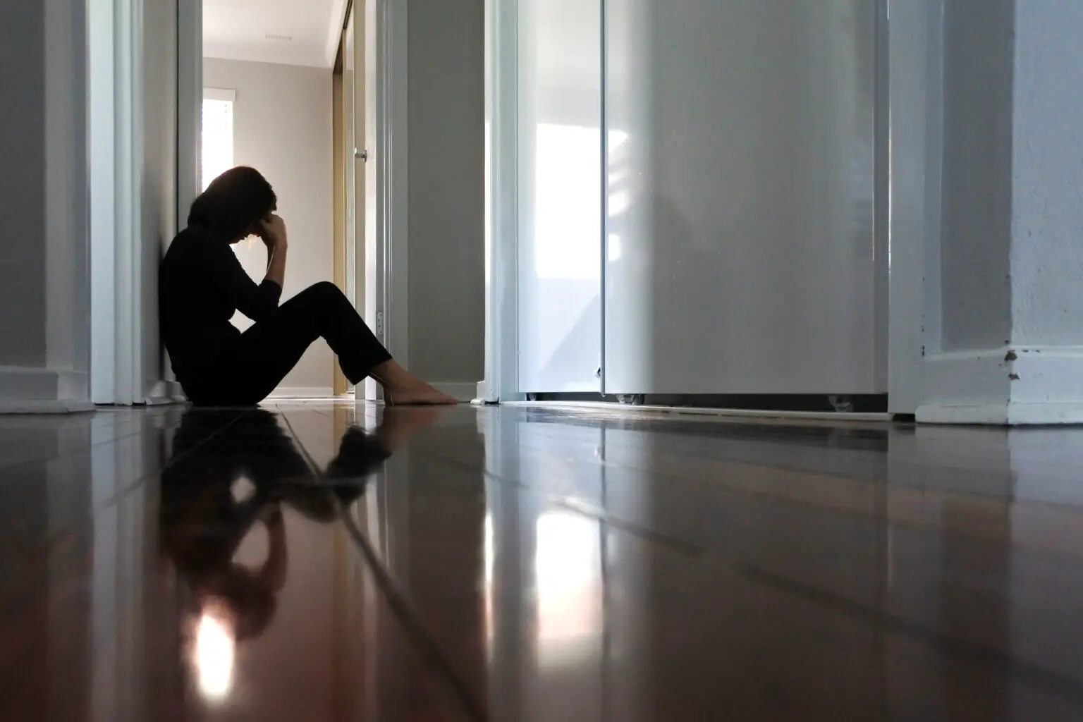 Sad adult woman sitting on dark home corridor floor.